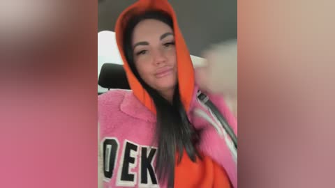 Video of a young woman with long black hair, wearing a bright pink hoodie with orange hood and \"DEEK\" partially visible, smiling confidently. Background is blurred.