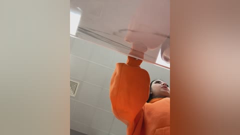 Video of a person in an orange jumpsuit climbing a white, reflective ceiling. The background features a white tiled wall with a ventilation grate. The image captures an overhead view, highlighting the contrast between the subject's bright attire and the muted surroundings.