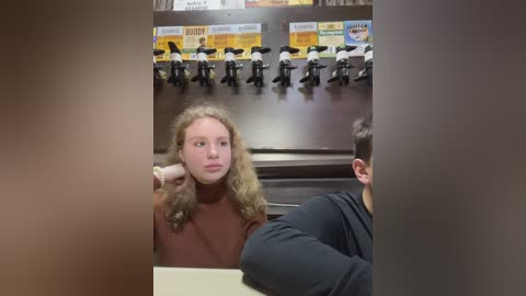 Media: A video of a young woman with long, curly blonde hair, wearing a brown turtleneck, sitting at a coffee counter, with several coffee cups and a menu board in the background.