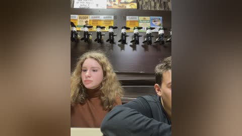 Media: Video of a young woman with curly blonde hair in a brown turtleneck sweater, seated at a dark wood table in a dimly lit cafe. Behind her, a row of toy soldiers and a menu board are visible.