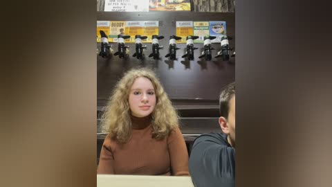 Media: Video of a young woman with curly blonde hair, wearing a brown turtleneck, seated in a diner with a dark wooden wall featuring miniature black and white figurines and \"Buddy\" signage.