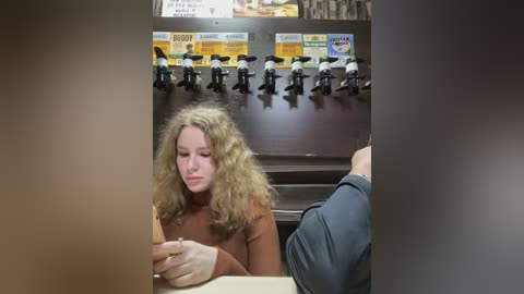 Media: A video of a young woman with curly blonde hair, wearing a brown sweater, sitting at a coffee shop counter, looking at her phone. Behind her, a row of toy figures and drink labels are displayed.