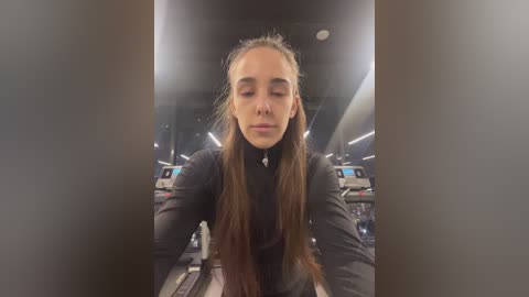 Media: Video of a young woman with long brown hair in a black turtleneck, eyes closed, standing in a dimly lit gym with exercise machines in the background.