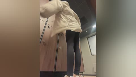 Media: Video of a person in a beige jacket and black leggings leaning against a beige locker in a dimly lit, institutional setting.