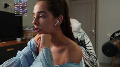 Media: Video of a young woman with light skin, dark hair, and red lipstick, wearing an off-shoulder light blue top, listening to white earbuds, in a modern bedroom with a bed and TV.