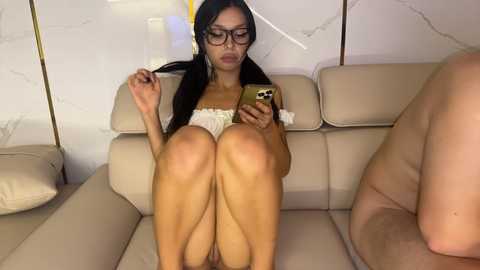 Media: Video of a dark-haired, bespectacled woman in a white, strapless top, sitting on a beige couch, texting on a gold phone, while a nude man reclines beside her.