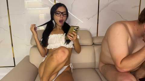 Media: Video of a young woman with long black hair, glasses, white lingerie, and a smartphone, sitting on a beige couch. A shirtless man with a muscular build sits next to her, partially visible. Marble walls and stairs are in the background.