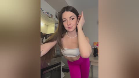Media: A video of a young woman with long brown hair, wearing a white crop top and bright pink leggings, posing in a bedroom with a mirror and various objects in the background.