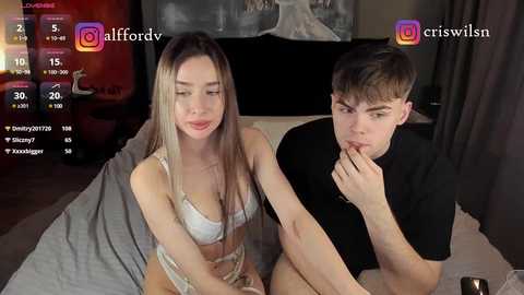 Media: Video of a young blonde woman in lingerie, and a young man in a t-shirt, sitting on a bed, in a dimly lit room with a gray blanket.