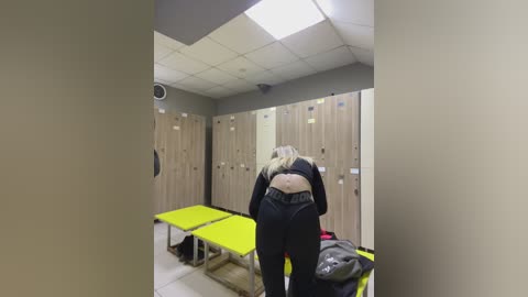 Media: Video of a blonde woman with fair skin in a gym locker room, wearing black workout gear, standing in front of beige wooden lockers with a bright yellow bench and a black bag on the floor.