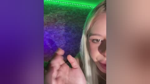 A close-up video of a blonde woman with light skin, partially obscured by a hand, showing her right eye and cheek. The background is dimly lit with green and purple hues, creating a moody atmosphere.