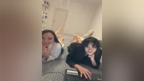 Video of two young women, one with glasses, lying on a bed in a sparsely furnished room. One wears a white shirt, the other a black top.