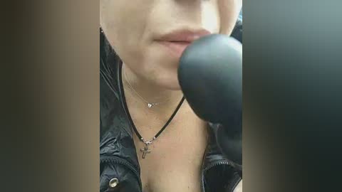 A close-up video of a woman's face, wearing a black leather jacket, necklace, and dark lipstick, with a microphone in focus, capturing a candid, intimate moment.
