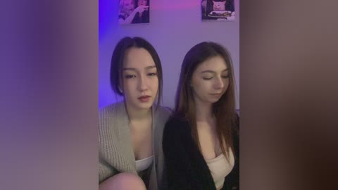 Media: Video of two young women, one with light skin and dark hair in a gray cardigan, the other with fair skin and brown hair in a black top, sitting in a dimly lit room with purple lighting.