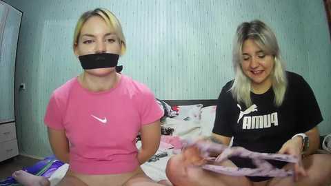 Media: Video of two women in a room: blonde with pink shirt, black tape over mouth; brunette in black shirt, holding pink underwear.