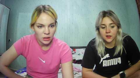 Media: Video of two young women with fair skin and blonde hair, wearing casual pink and black Puma shirts, sitting on a bed with floral sheets in a dimly lit room with vertical striped wallpaper.