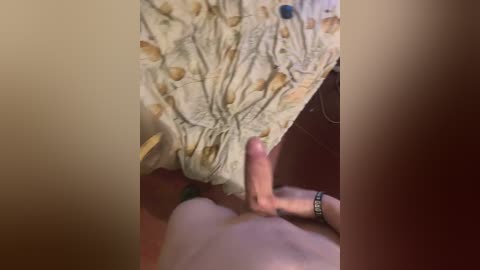 A close-up video captures a man's erect penis, partially covered by a patterned blanket, with a wristwatch on his left hand. The background is dimly lit, focusing on the intimate moment.
