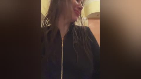 Media: Video of a woman with wet hair, wearing a black jacket, smiling slightly, blurred background with a yellow lampshade.