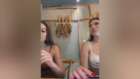 Media: Video of two young women with fair skin and long brown hair, wearing white tank tops, standing in a rustic room with wicker panels and dried wheat bundles on a blue wall.