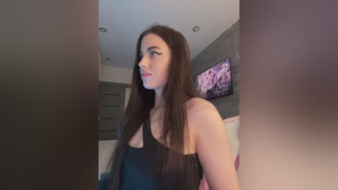 Video of a young woman with long, straight brown hair, wearing a black, spaghetti-strap top, standing in a modern bedroom with a gray wall and a pink floral painting.