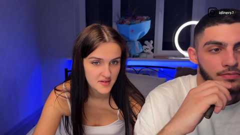 Video of a young Caucasian woman with long, straight brown hair and fair skin, wearing a white tank top, sitting closely to a dark-skinned man with a trimmed beard and mustache. Background features a nightstand with a lit ring light and a decorative bowl.