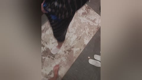 Video of a person with light skin, wearing a black and white patterned shirt, standing on a beige and gray tiled floor, with a pair of white slippers near their feet.