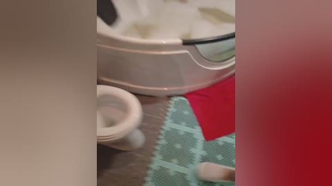 A blurry video captures a bathroom scene with a white toilet and a green and red bath mat on a gray tiled floor. A person's foot is visible in the bottom right corner.