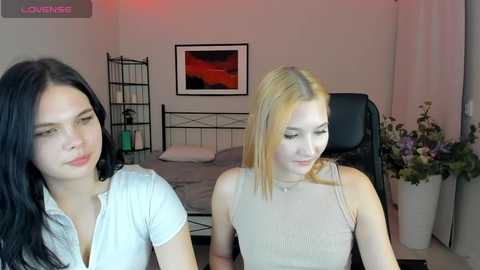 Video of two women in a modern bedroom: one with long black hair in a white top, the other with blonde hair in a beige top, sitting at a desk.
