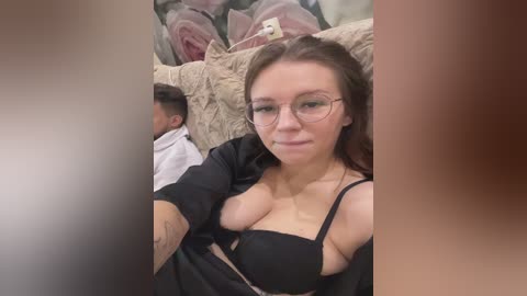 Media: Video of a young woman with fair skin, glasses, and a black bra, seated on a beige couch with a blanket, next to a sleeping baby.