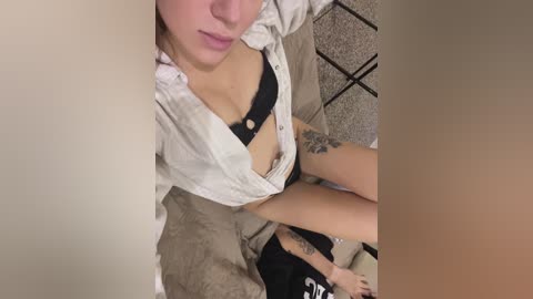 Media: Video of a fair-skinned woman with a black bra, sitting on a beige carpet, partially obscured by a beige curtain.