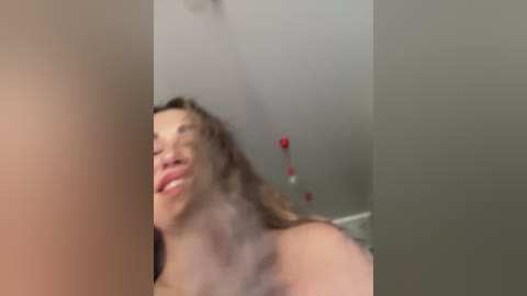 A blurry, candid video of a smiling, nude woman with light skin, shoulder-length curly brown hair, and a playful expression. Background features a white wall with a red heart-shaped decoration.