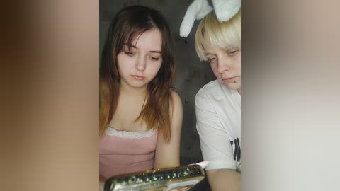 Media: Video of two young women, one with brown hair and bunny ears, the other with blonde hair, in a dimly lit room, wearing casual clothing, with a vintage camera in focus.