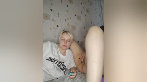 Media: A video of a young, fair-skinned, blonde woman with short hair, wearing a white T-shirt and no pants, lying on a bed with a patterned wallpaper background, eyes closed, holding a blue object.