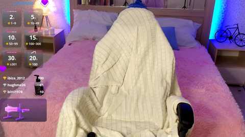 Video of a person, head covered, lying on a pink bed with white pillows, wearing a beige knitted blanket. A digital screen displays a social media feed with heart and message icons.