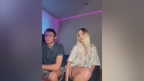 Media: Video of a young man with short brown hair and a young woman with long blonde hair, both sitting on a couch. They have serious expressions, in a dimly lit room with purple LED strip lighting.