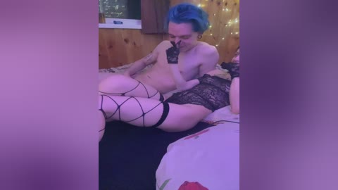 Video of a pale-skinned, blue-haired transgender woman with tattoos, wearing black lace lingerie and fishnet stockings, lying on a bed in a dimly lit room with fairy lights and a window.
