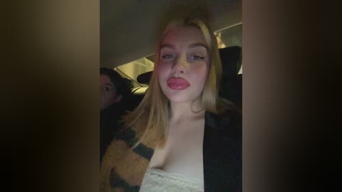 Video of a blonde woman with pink lipstick, wearing a black jacket over a beige top, seated in a dimly-lit car.