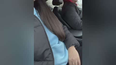 Media: A video showing a person with long brown hair wearing a blue shirt and black jacket seated in a car, next to another person in a black jacket.