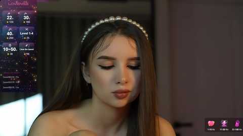 Media: A video shows a young woman with long brown hair, wearing a sparkling headband, looking down thoughtfully. The background is blurred, displaying a dimly lit room. A digital overlay displays her live stream stats.