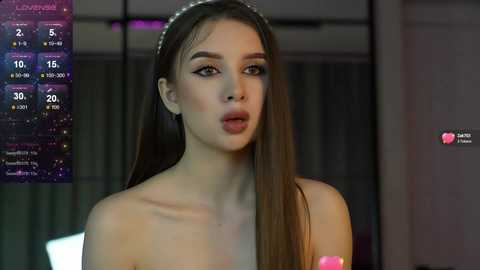 Media: A video of a young Caucasian woman with long brown hair and a sparkling headband, wearing minimal makeup, lips slightly parted, set against a dimly lit, modern background with a TV weather forecast visible.