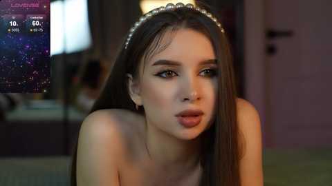 Media: Video of a young woman with long brown hair, wearing a pearl headband, and dark eyeliner, indoors, blurred background, looking pensive.