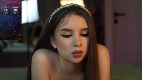 Media: Video of a young woman with long brown hair, wearing a pearl headband, and makeup. She's in a dimly lit room, with a digital clock displaying \"12:06\" in the background.