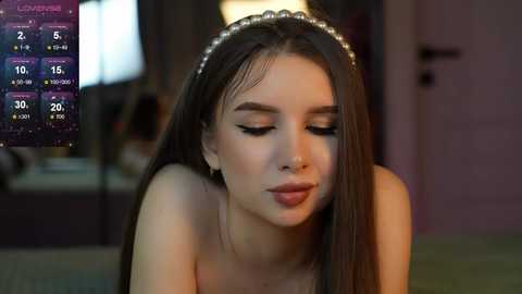 Media: Video of a young woman with long brown hair, wearing a pearl headband, looking down. Background shows a dimly lit room with a TV displaying weather information.