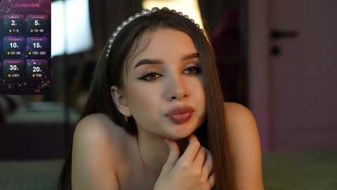 Media: Video of a young woman with long brown hair, wearing a headband and makeup, indoors with a dimly lit background and a weather forecast overlay.