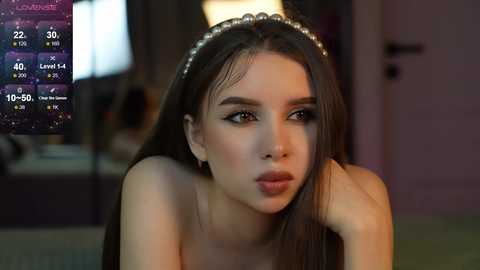 Media: Video of a young woman with long brown hair, wearing a pearl headband, looking pensive. Background shows a dimly lit room with a TV screen displaying weather information.