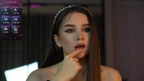 Media: Video of a young woman with long brown hair, wearing a headband, looking thoughtful, finger to her lips, in a dimly lit room.