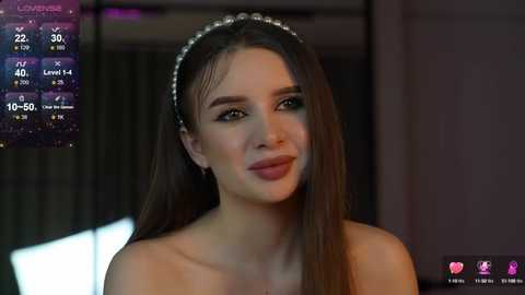 Media: Video of a fair-skinned woman with long brown hair, wearing a sparkling headband, smiling slightly. Background shows a dimly lit room with a digital display and a blurred window.