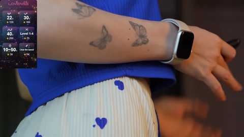 Media: Video of a person with fair skin, wearing a blue sleeveless top and white pants with blue butterflies, showcasing a smartwatch on their left wrist.