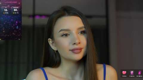 Media: Video of a young woman with straight, dark brown hair and fair skin, wearing a blue sleeveless top, indoors with a blurred background featuring a digital interface displaying a starry night sky.