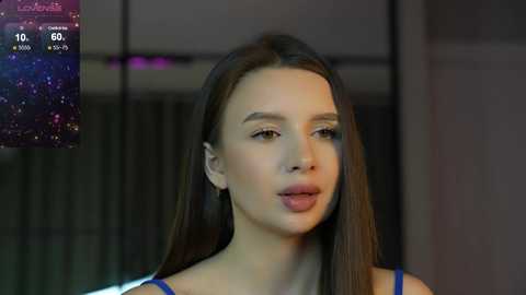 Media: Video of a young woman with long brown hair, wearing a blue top, set in a modern, dimly lit room with a digital clock displaying 10:06.